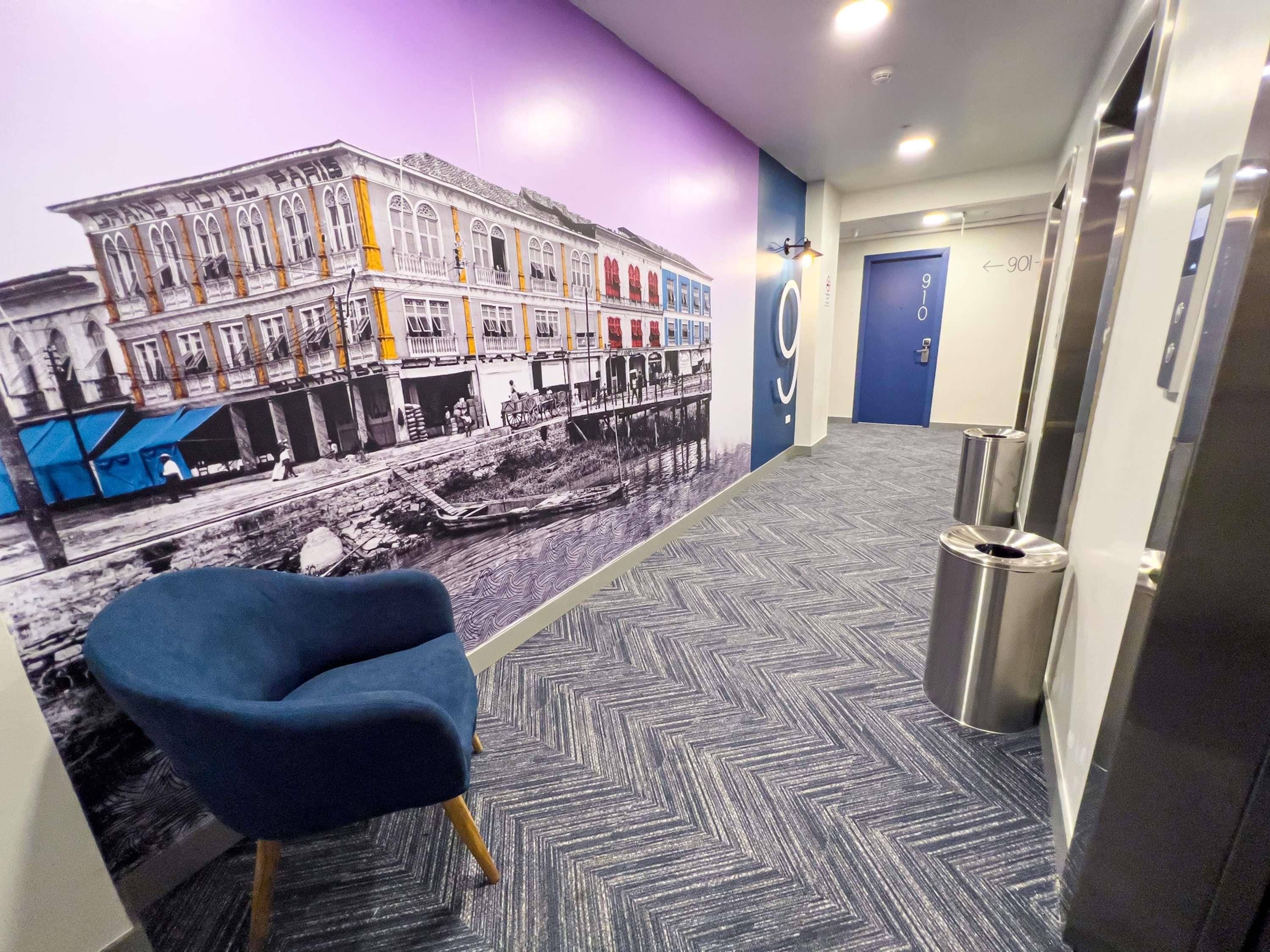 Un couloir orné d’une fresque représentant un paysage urbain, avec une chaise bleue et une poubelle.