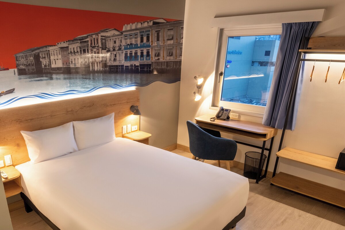 Habitación de hotel con cama, escritorio, silla y vistas a la ciudad por la ventana.
