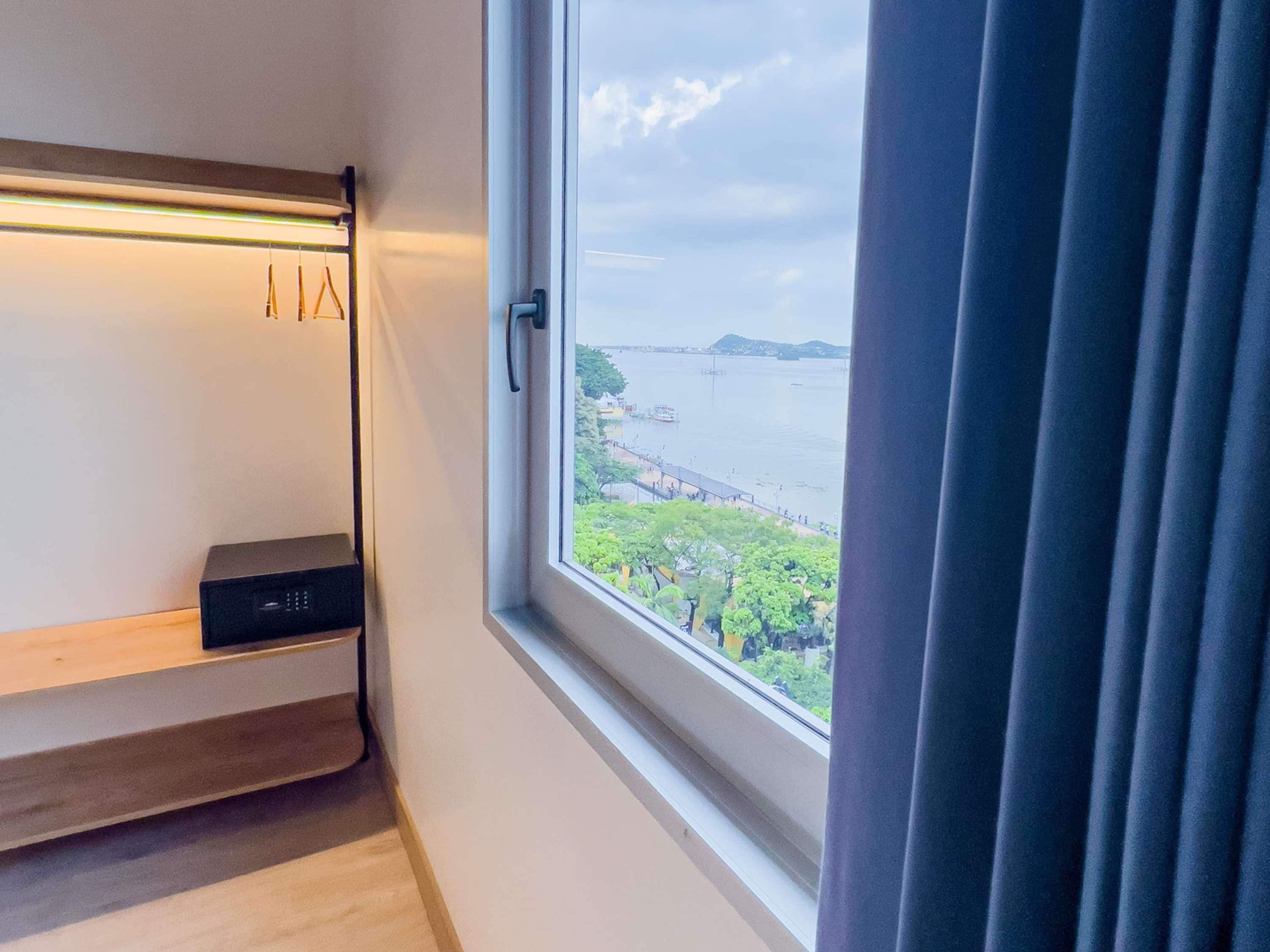 Una habitación con ventana que da a un cuerpo de agua, una estantería con un dispositivo negro y una cortina entreabierta.