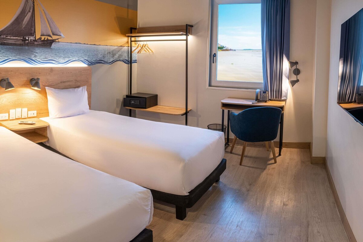 Een hotelkamer met twee bedden, een bureau, een stoel, een nachtkastje met een lamp, een klein plankje en een raam met uitzicht op zee.