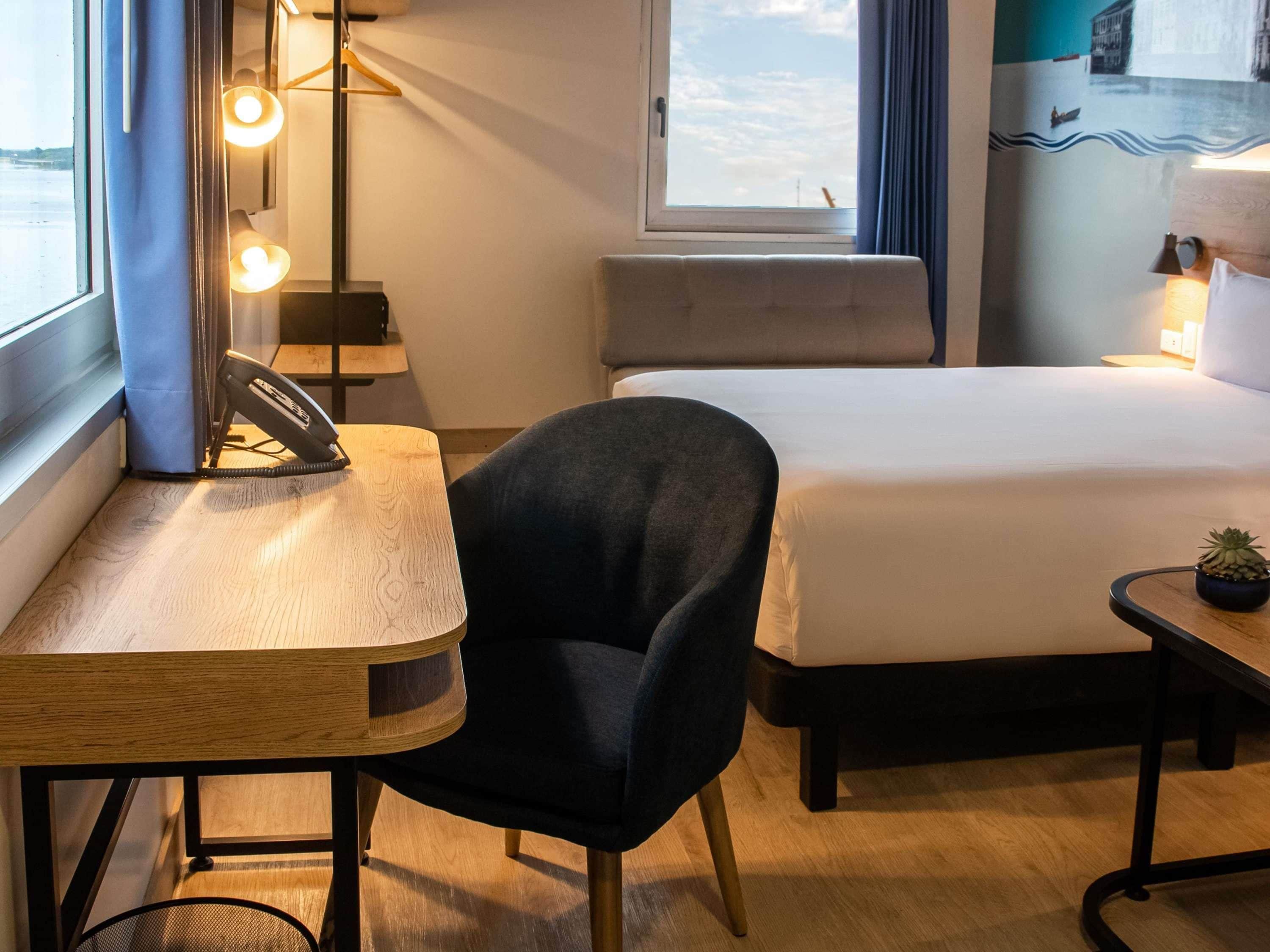 Ein Hotelzimmer mit Bett, Schreibtisch mit Stuhl, einem kleinen Tisch mit Pflanze und einem Fenster mit Blick.