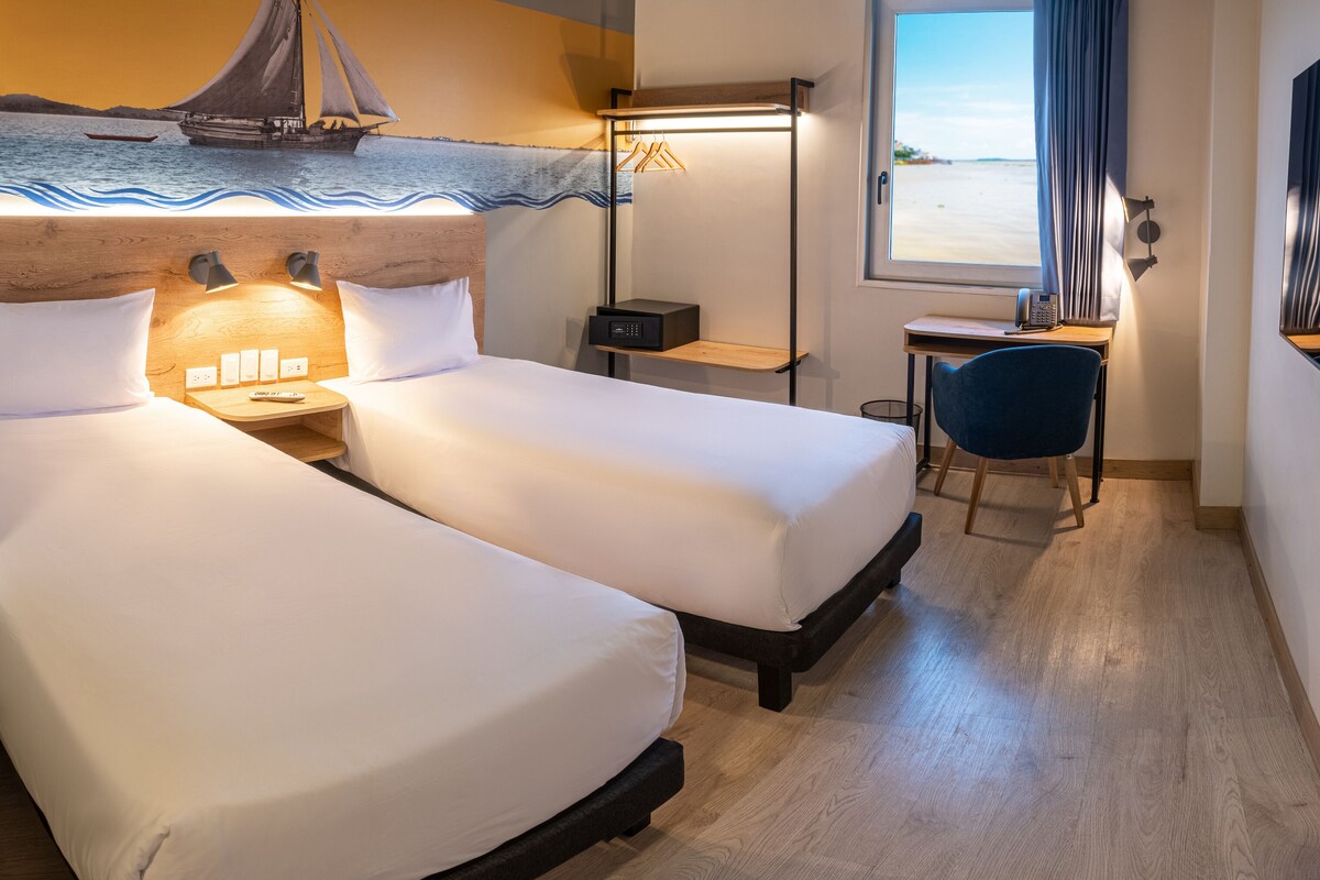 Habitación de hotel con dos camas, un escritorio, una silla, una mesita, un televisor montado en la pared y una ventana con vista a la playa.