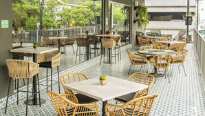 Een terras met rieten stoelen en tafels, een vloer met patroon en uitzicht op groen door grote ramen.