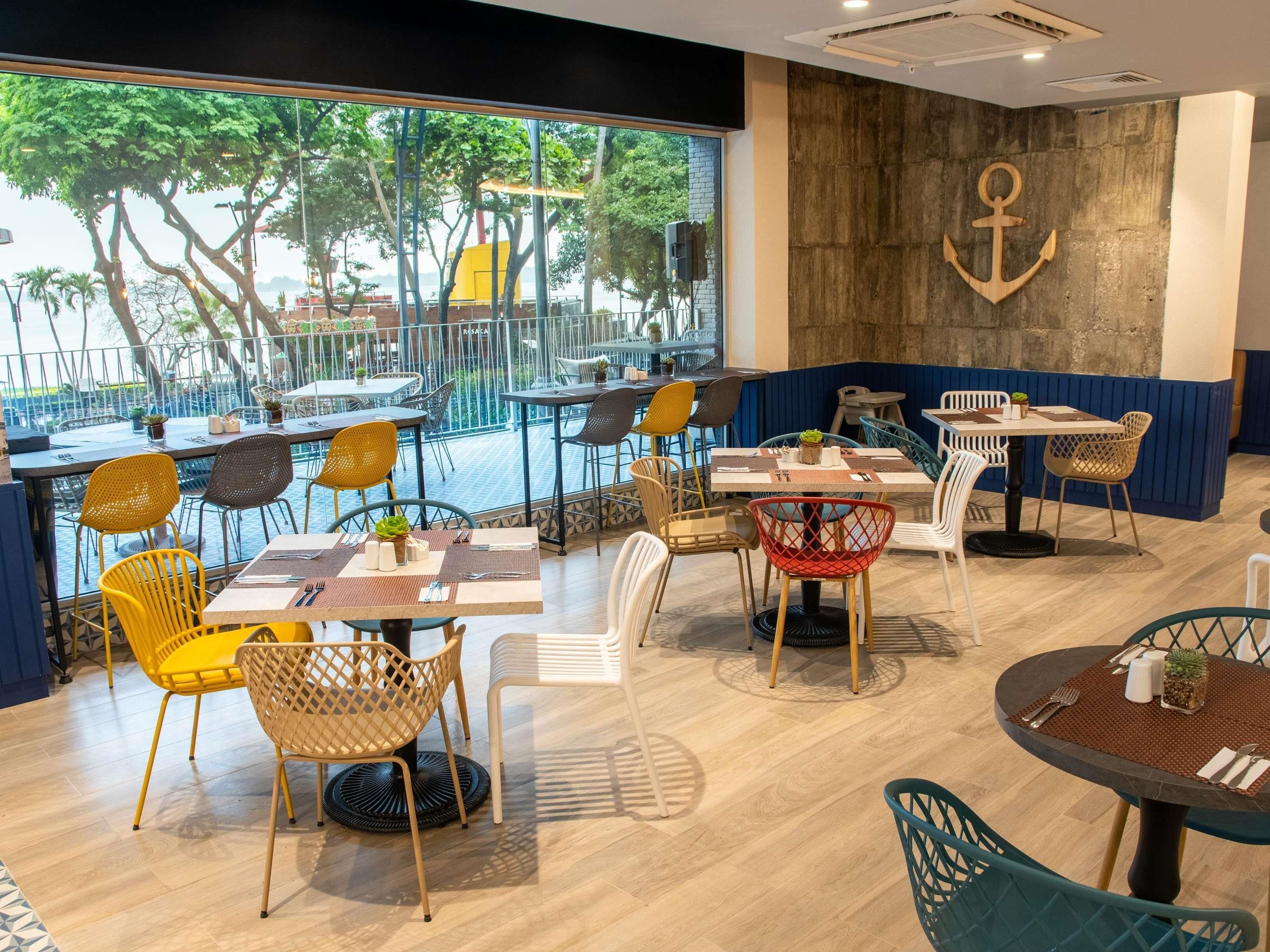 Un comedor interior con mesas y sillas, un ventanal grande que da a una terraza con asientos exteriores, y una pared decorada con un ancla.