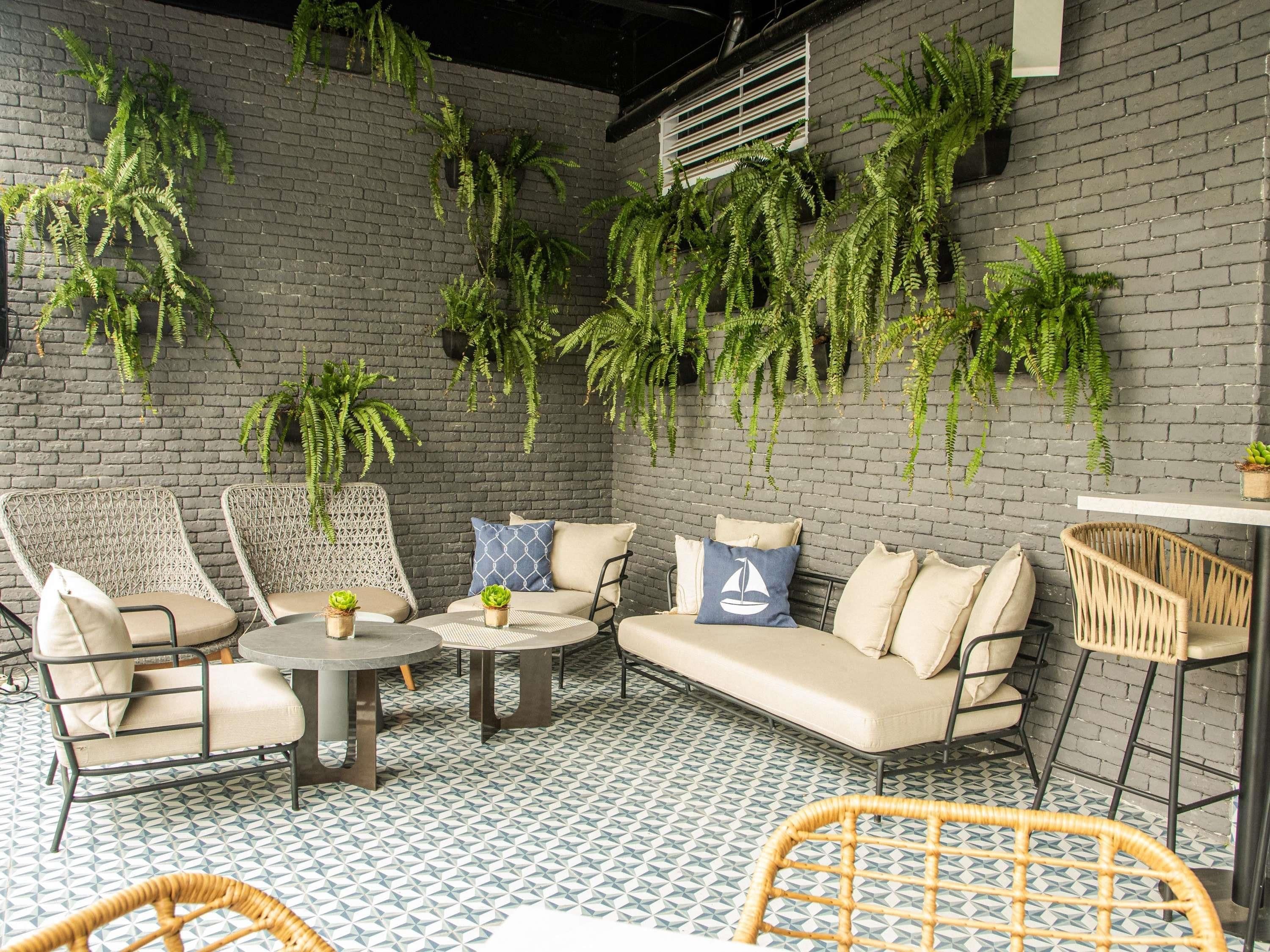 Un rincón al aire libre acogedor con muebles de mimbre, piso estampado y plantas verdes exuberantes frente a un muro de ladrillo.