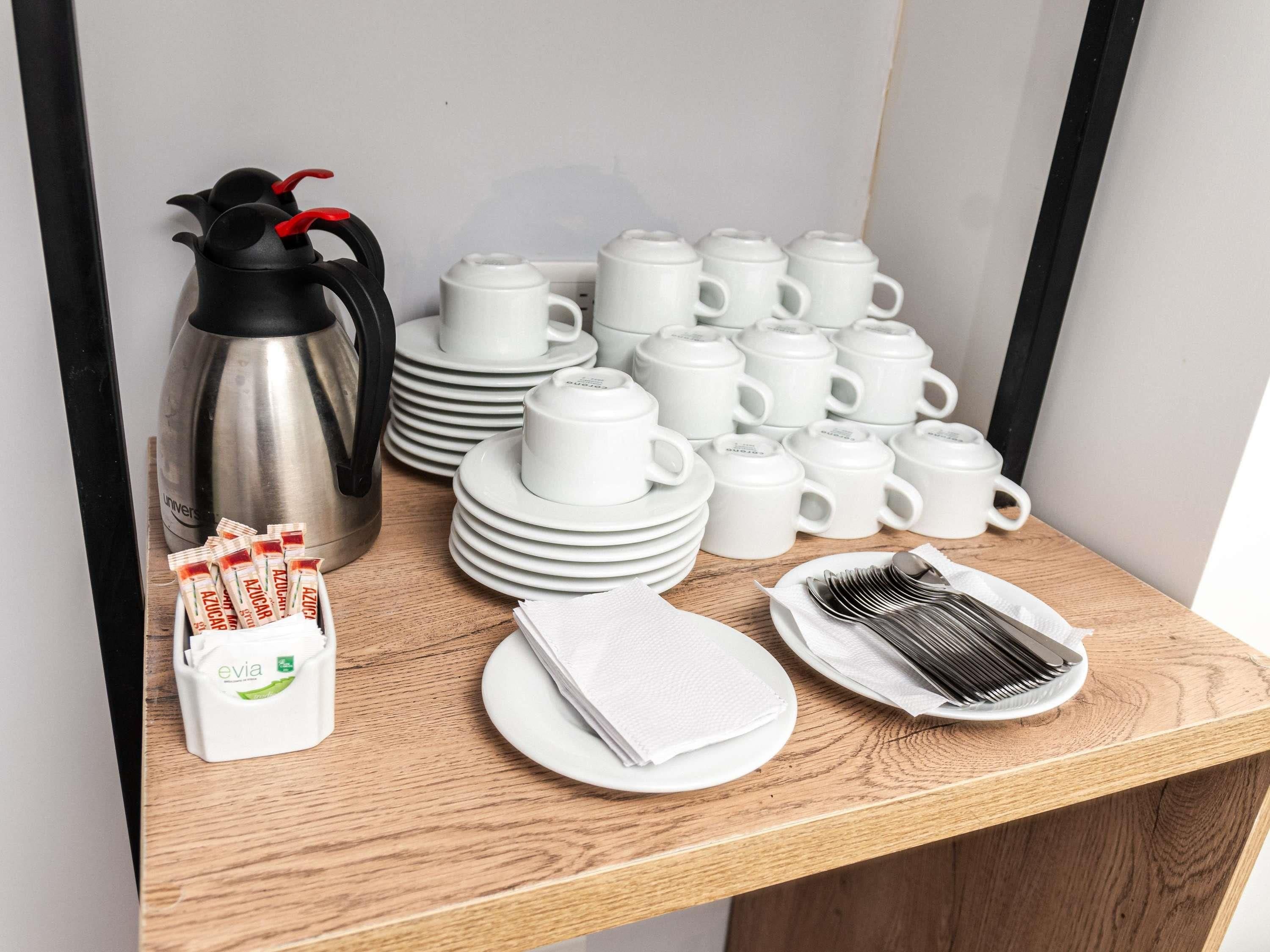 Uma mesa de madeira com uma composição organizada para o café da manhã, incluindo uma cafeteira, vários copos e pratos, e utensílios.