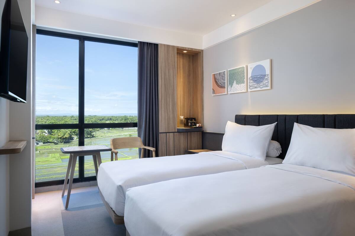 A hotel room with two beds, a desk, and a chair. There is a large window offering a view of greenery.