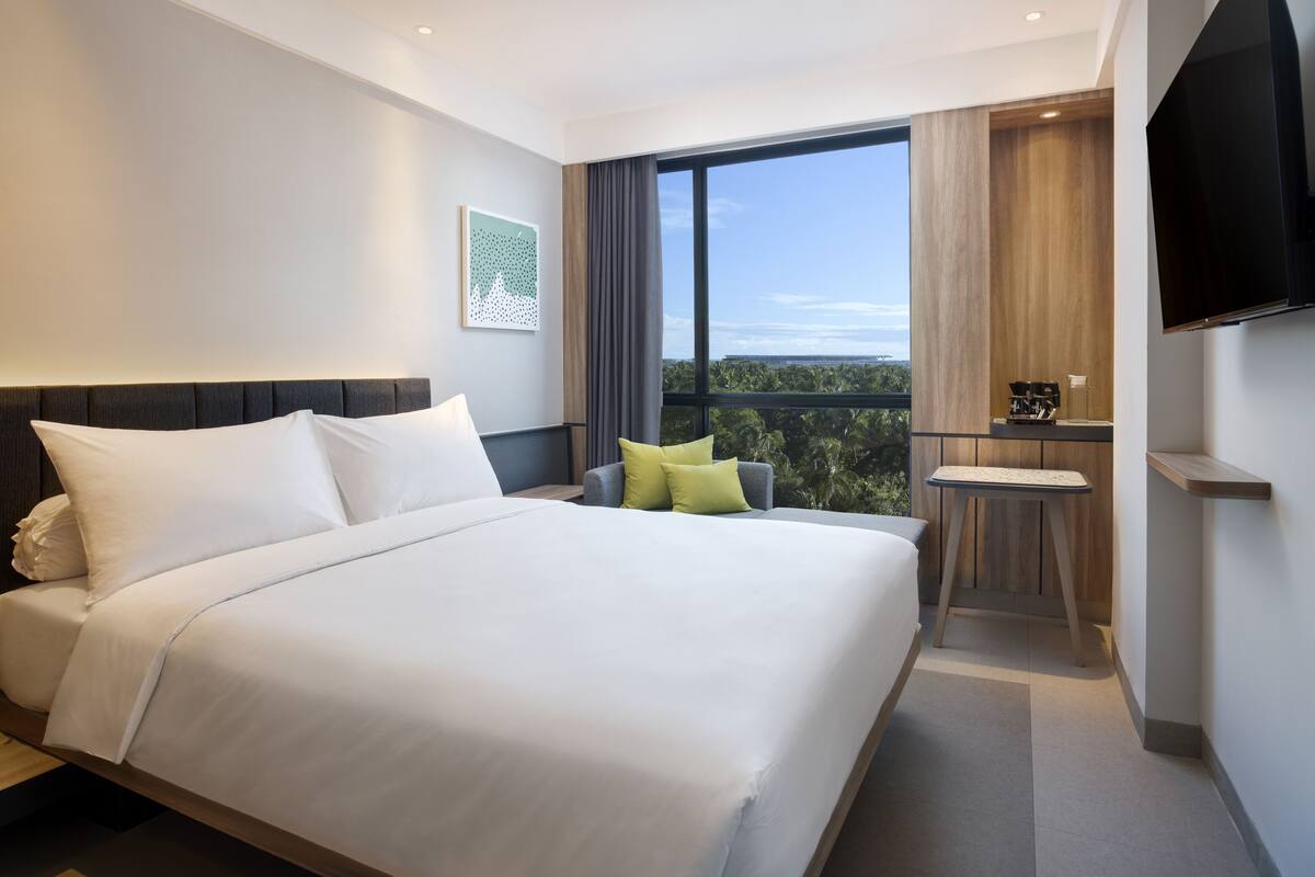 A modern hotel room with a large bed, a seating area with a chair and a small table, a flat-screen TV mounted on the wall, and a large window offering a view of the outdoors.