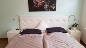 Een tweepersoonsbed met een gestoffeerd hoofdbord, twee nachtkastjes met lampen, en een bloemig kunstwerk erboven.