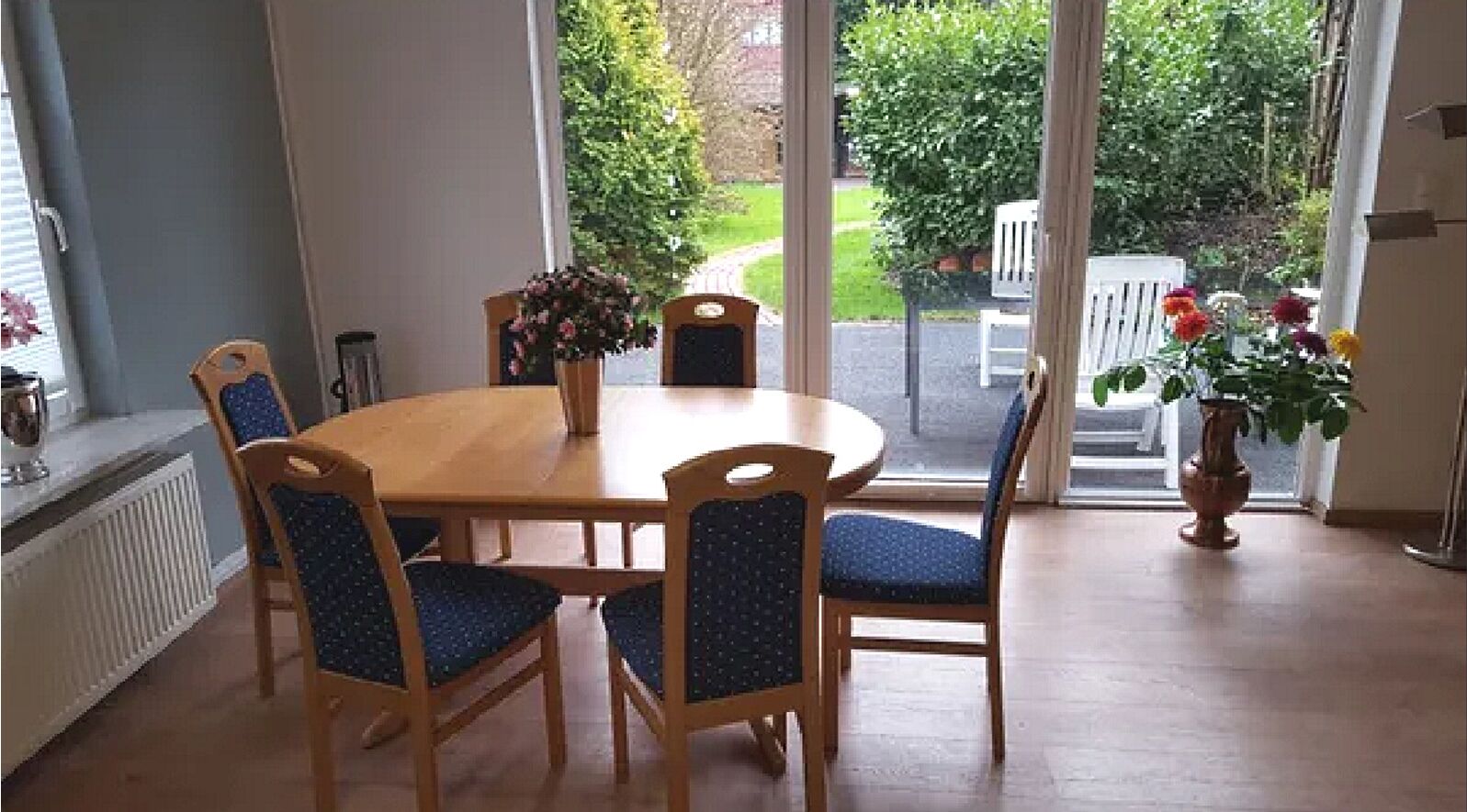 Ein Essbereich mit Holztisch und blau gepolsterten Stühlen, einer Blumenvase und Blick auf einen Garten durch Glastüren.