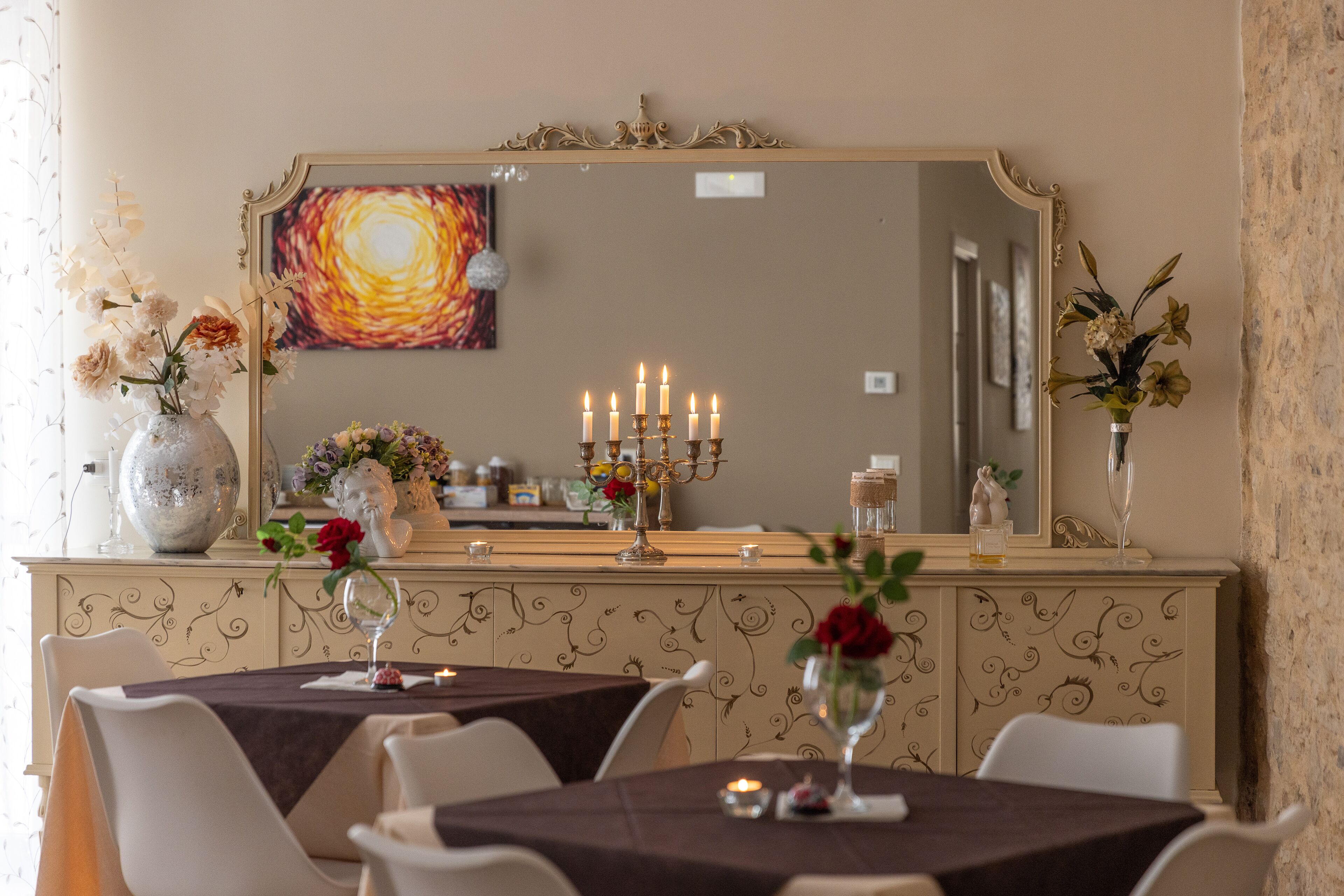 A dining area with tables set for two, a large mirror, and decorative vases with flowers.