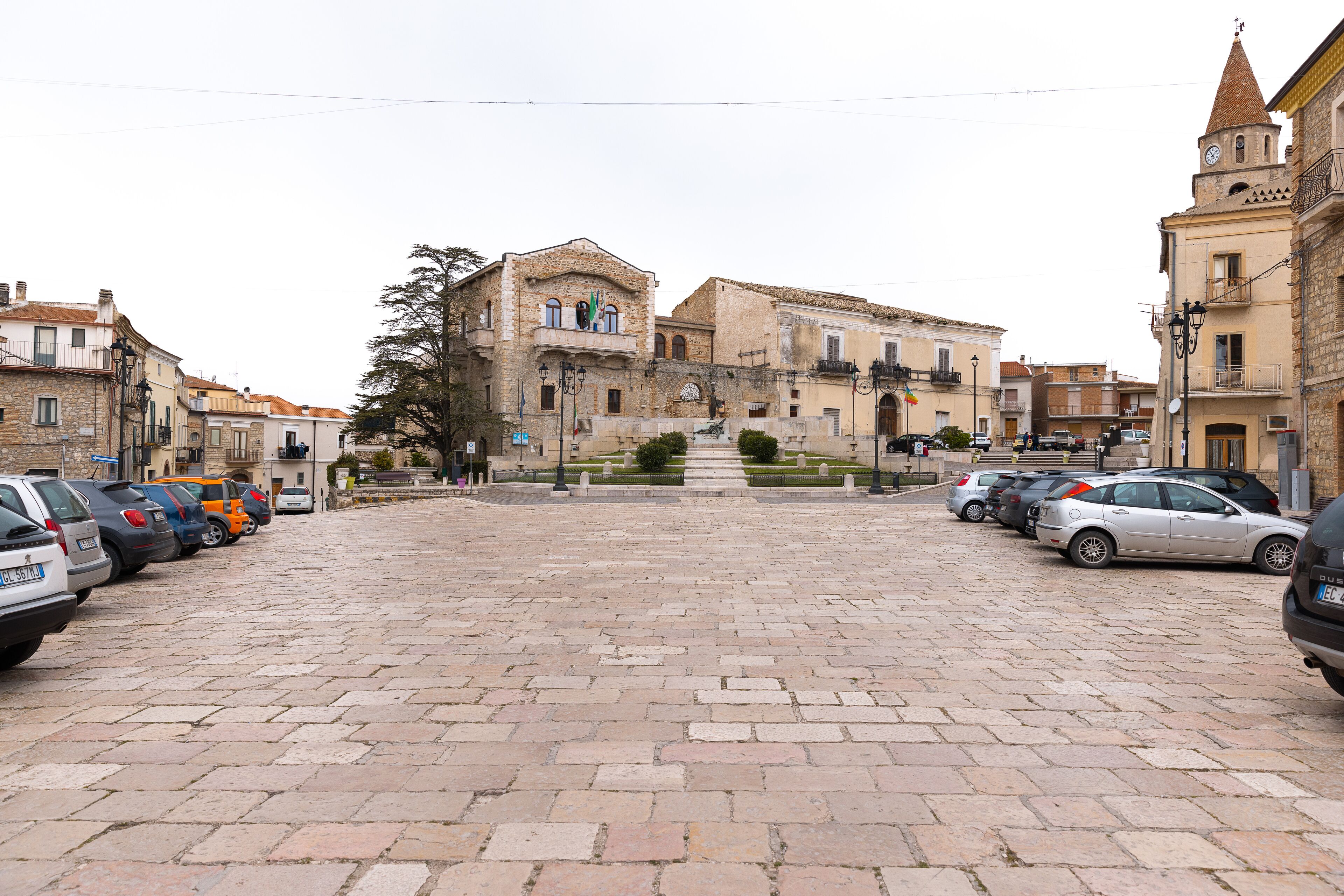 Una plaza pavimentada con coches aparcados y edificios históricos con balcones y una torre del reloj.