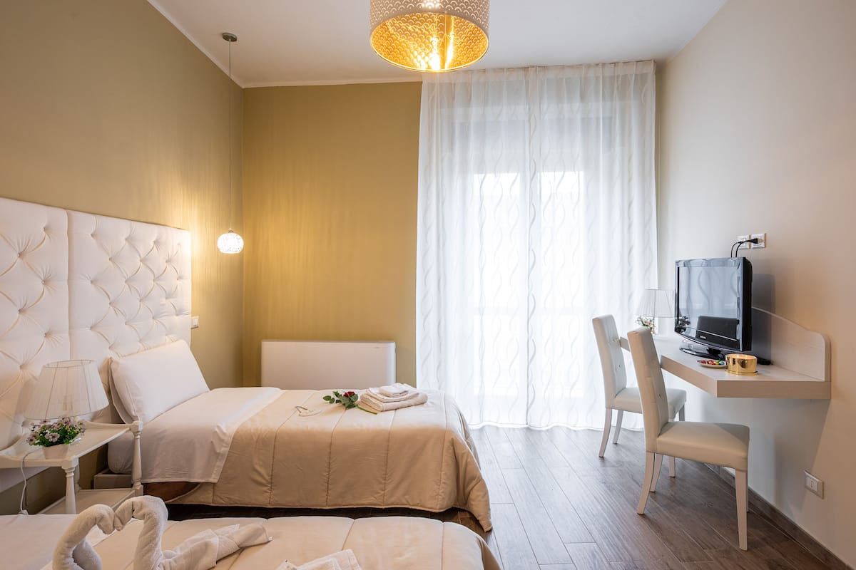A hotel room with two beds, a desk with a television, and a large window with sheer curtains.
