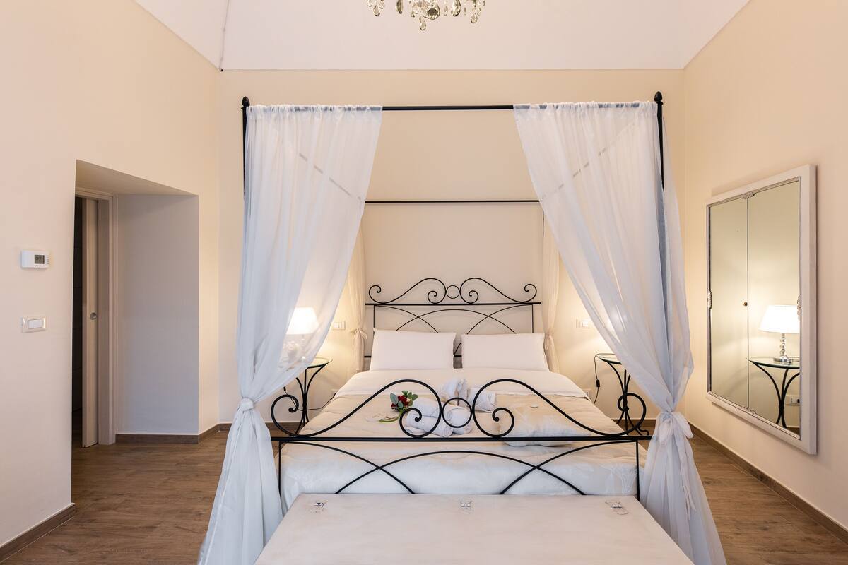 A four-poster bed with white linens, a bedside table with a lamp, and a large mirror on the wall.