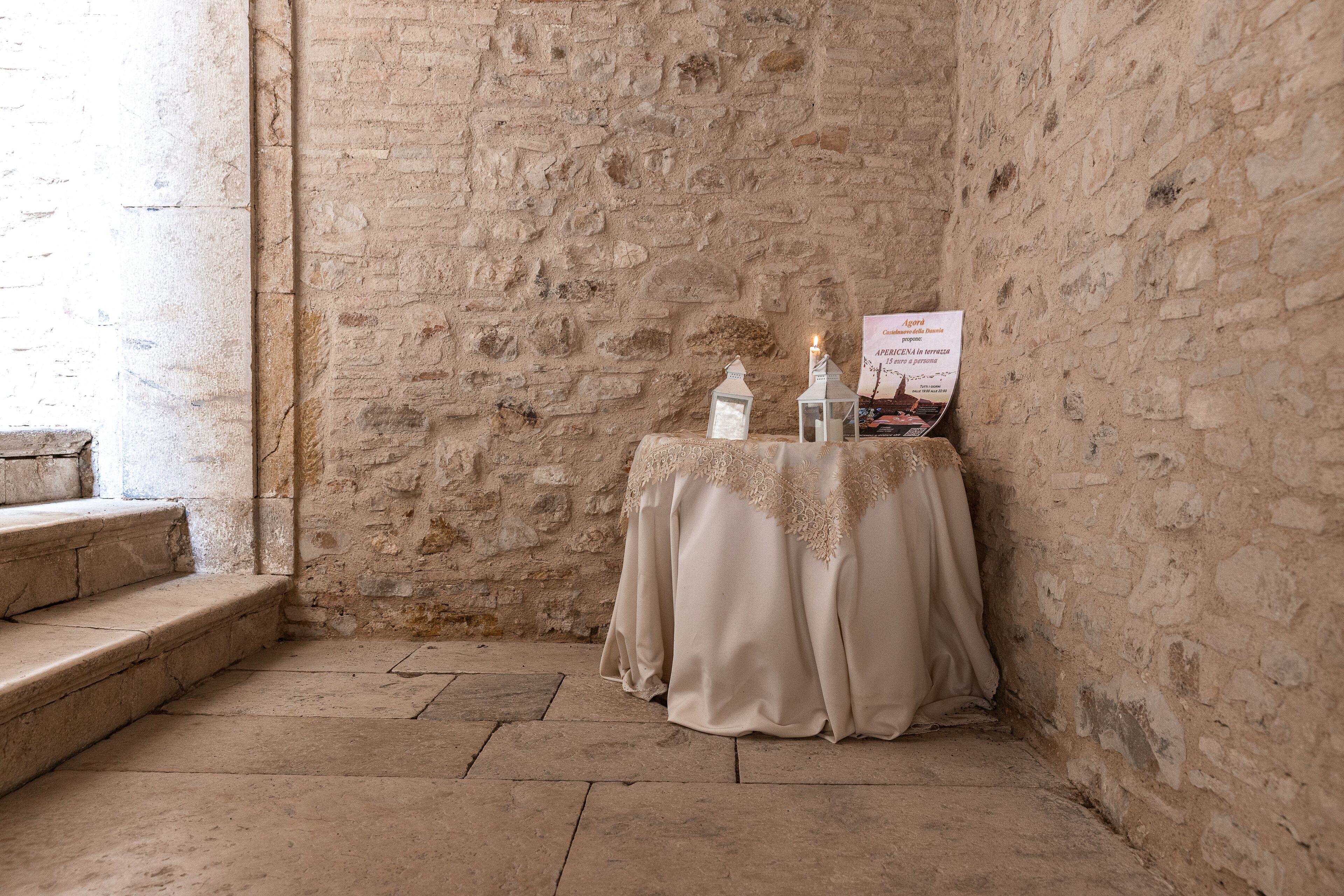 A stone-walled room with a table covered by a white cloth, two candles, and a sign.