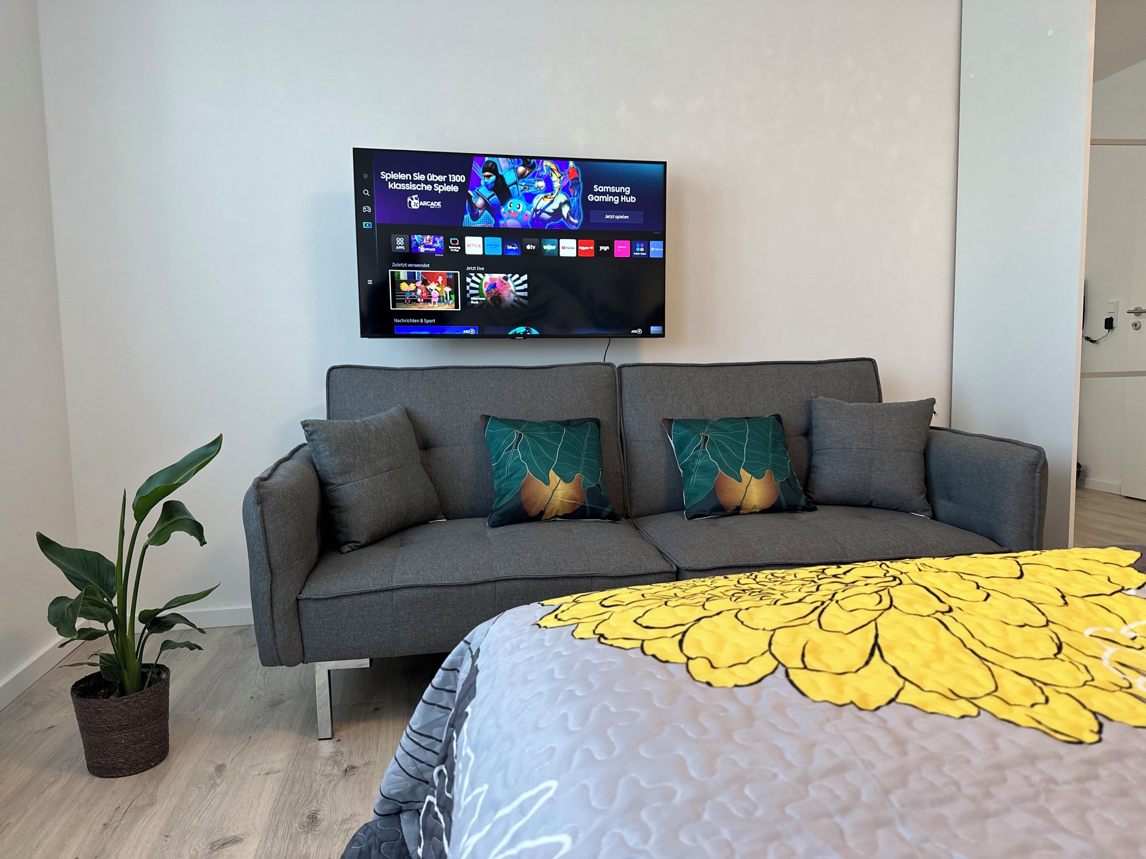 A modern living room with a grey sofa, two decorative pillows, a large flat-screen TV mounted on the wall, and a potted plant.