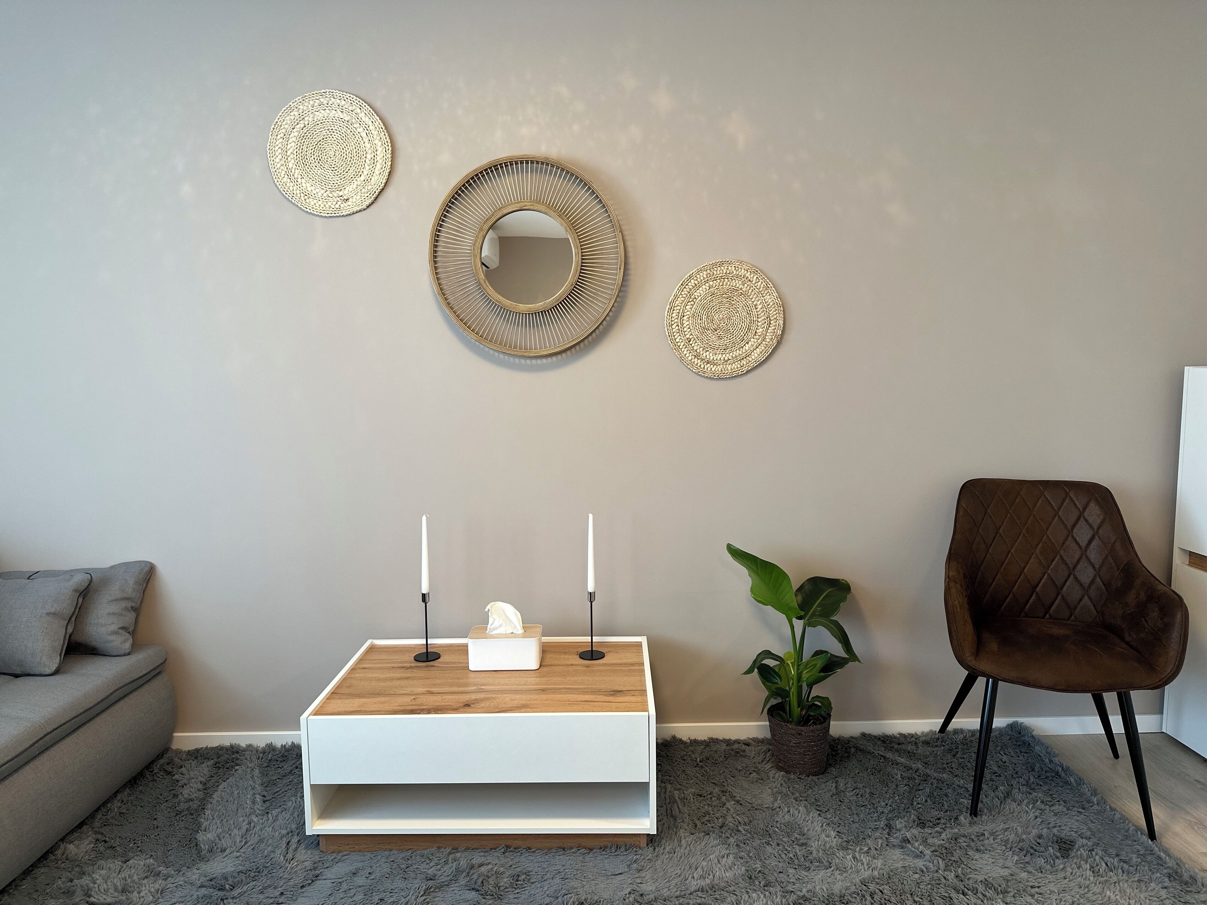 Een woonkamer met een grijze bank, een witte console tafel met kaarsen, een bruine fauteuil en decoratieve wanddecoratie.