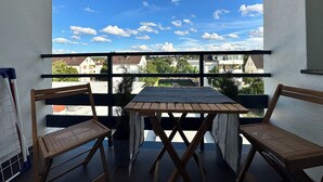 A balcony with a wooden table and two wicker chairs, overlooking a residential area with houses and trees.