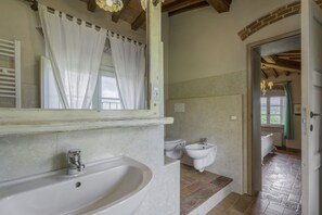 Un baño con un lavamanos grande, un bidé y un inodoro. Hay un espejo sobre el lavamanos y una ventana con cortinas.
