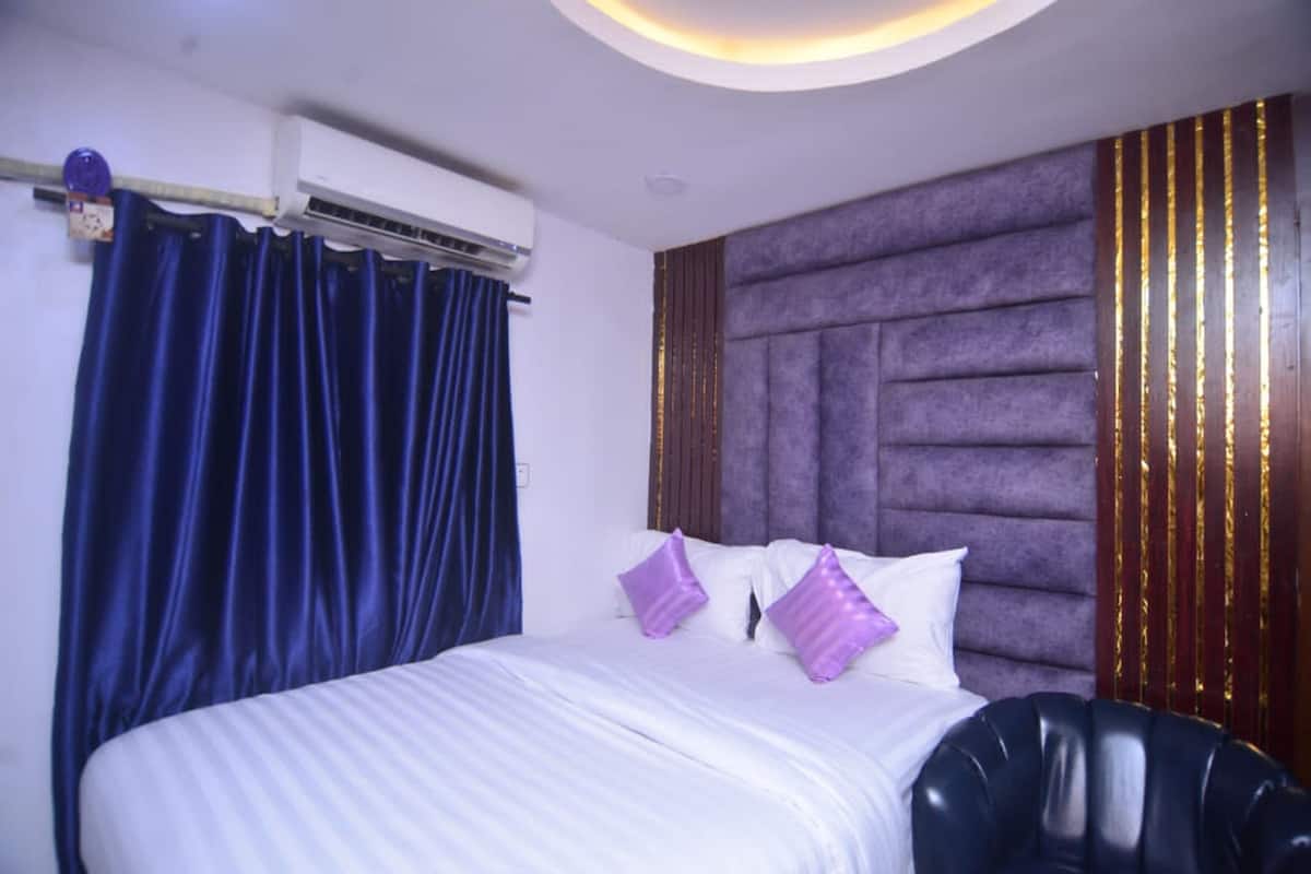 A hotel room with a bed, a headboard, a window with curtains, and an air conditioning unit.