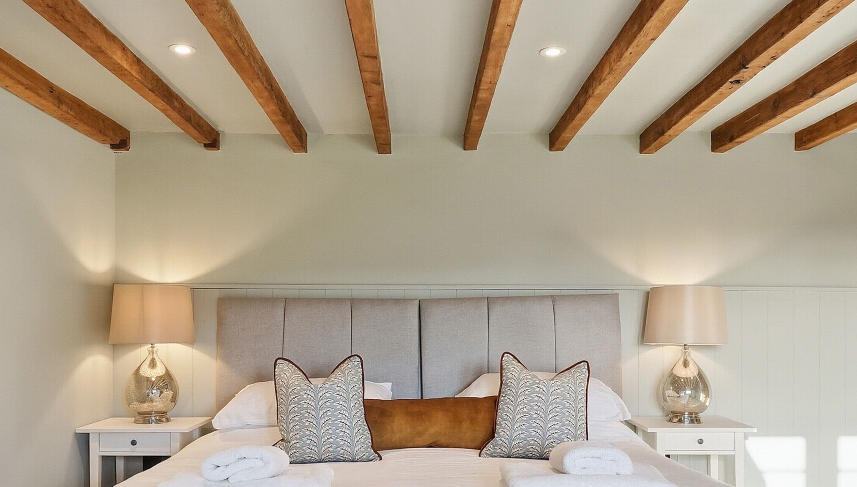 A bedroom with a wooden ceiling, a large bed with two pillows, two bedside tables with lamps, and a headboard.