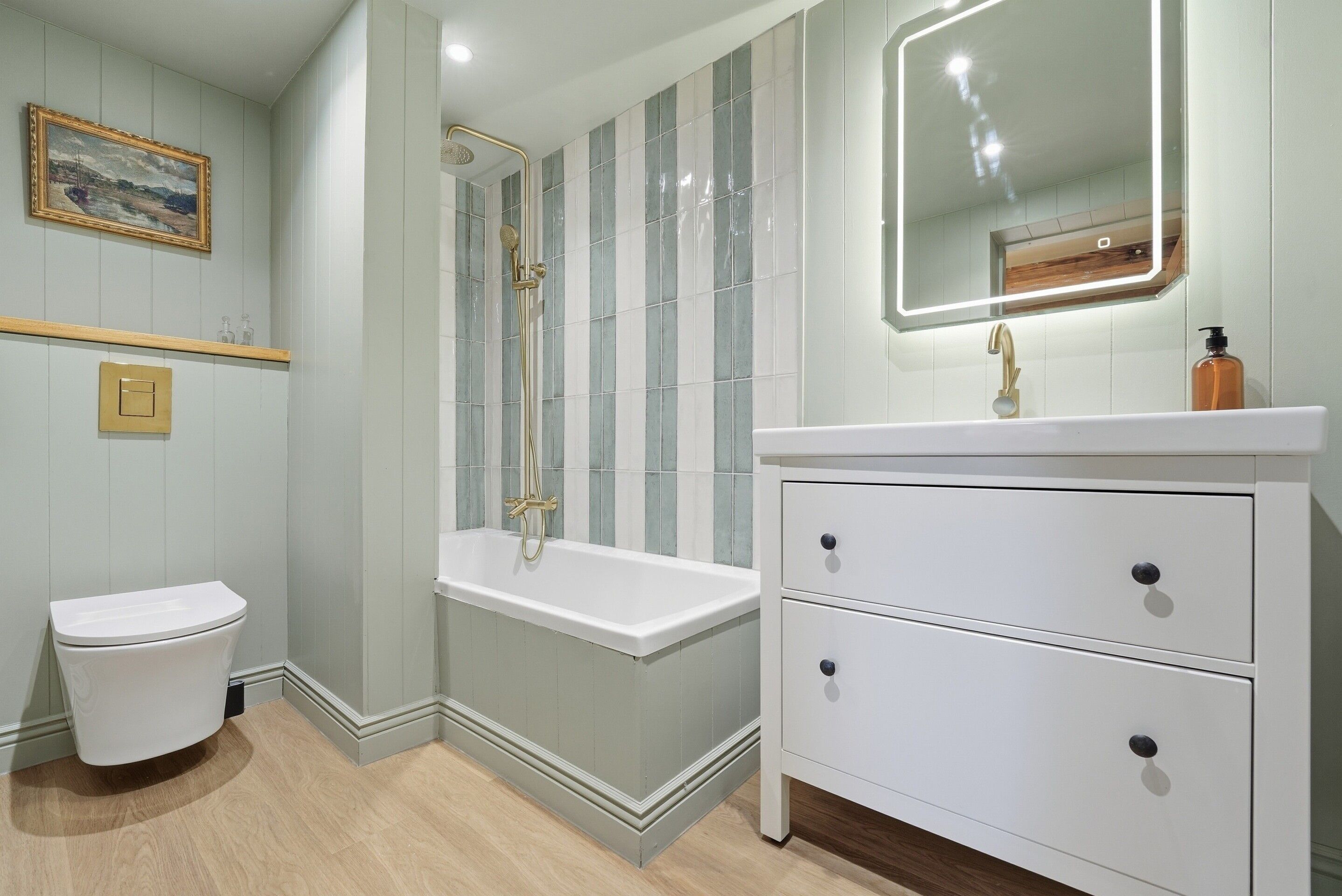 A bathroom with a white toilet, a bathtub, a vanity with drawers, a mirror, and a framed picture on the wall.