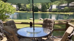 A round glass table with four cushioned chairs under a green umbrella, overlooking a calm lake with a wooden deck and surrounding greenery. - Fishing, skiing, golfing, water parks, restaurants, outlets  , family activity (Blakeslee)