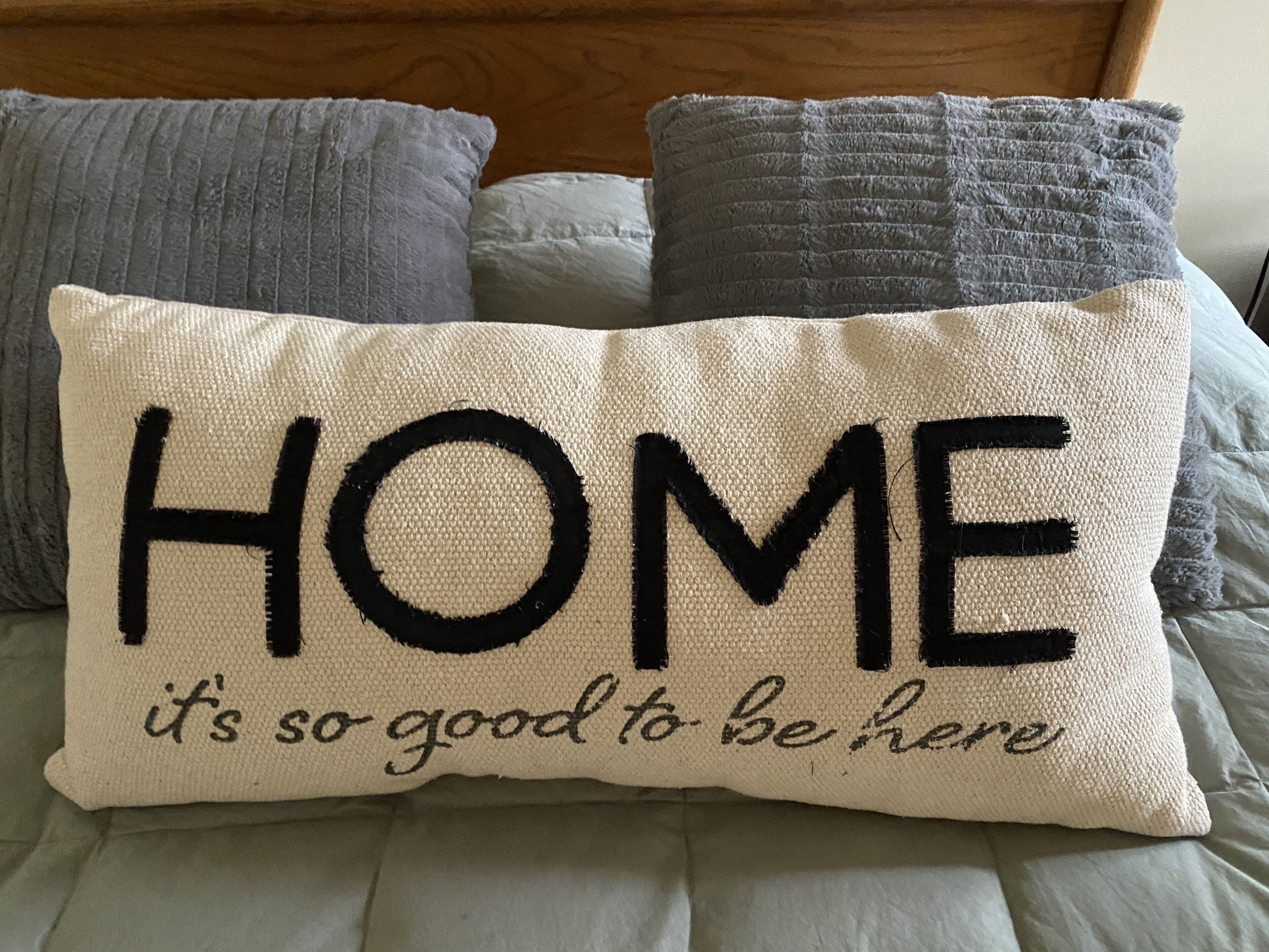 A pillow with the text "HOME it's so good to be here" on a bed with grey bedding.