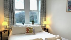 Cama bem arrumada com lençóis brancos, duas luminárias de cabeceira, travesseiro estampado e uma janela com cortinas.