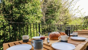 Uma mesa de madeira posta com pratos, canecas, um pão e um croissant numa varanda com guarda-corpo de metal e vegetação exuberante ao fundo.