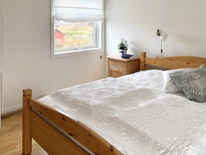 A bedroom with a wooden bed, a nightstand with a lamp, a window with blinds, and a view of a red building outside. - 4 Star Holiday Home in Odensbacken (Odensbacken)