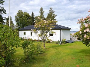 A white wooden house with a black roof, surrounded by a well-maintained lawn and flowering trees. - 4 Star Holiday Home in Odensbacken (Odensbacken)