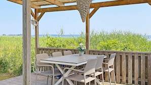 Una terraza exterior de madera con una mesa y sillas, una cesta de flores colgante, y vistas al mar y a los campos.