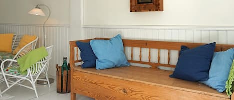 Un banc en bois avec des coussins bleus, une chaise en osier blanche avec des coussins jaunes, une bouteille verte et une horloge murale.