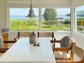 A dining area with a table and chairs, a hanging light fixture, and a view of a grassy landscape and water through large windows. - 4 Star Holiday Home in Odensbacken (Odensbacken)