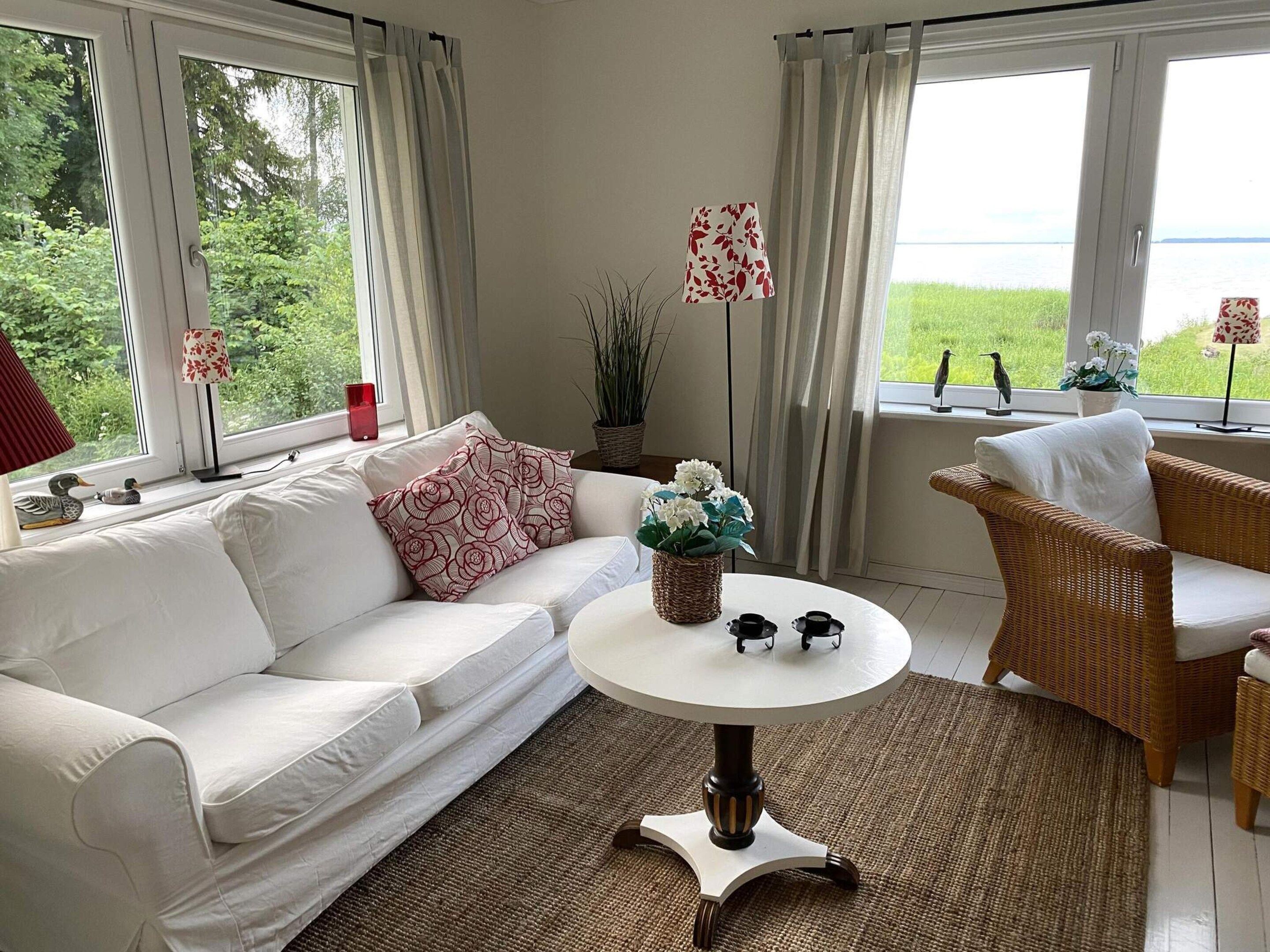 Una sala de estar con un sofá blanco, un sillón de mimbre, una mesa redonda con un jarrón de flores y vistas a la vegetación y al agua a través de grandes ventanales.