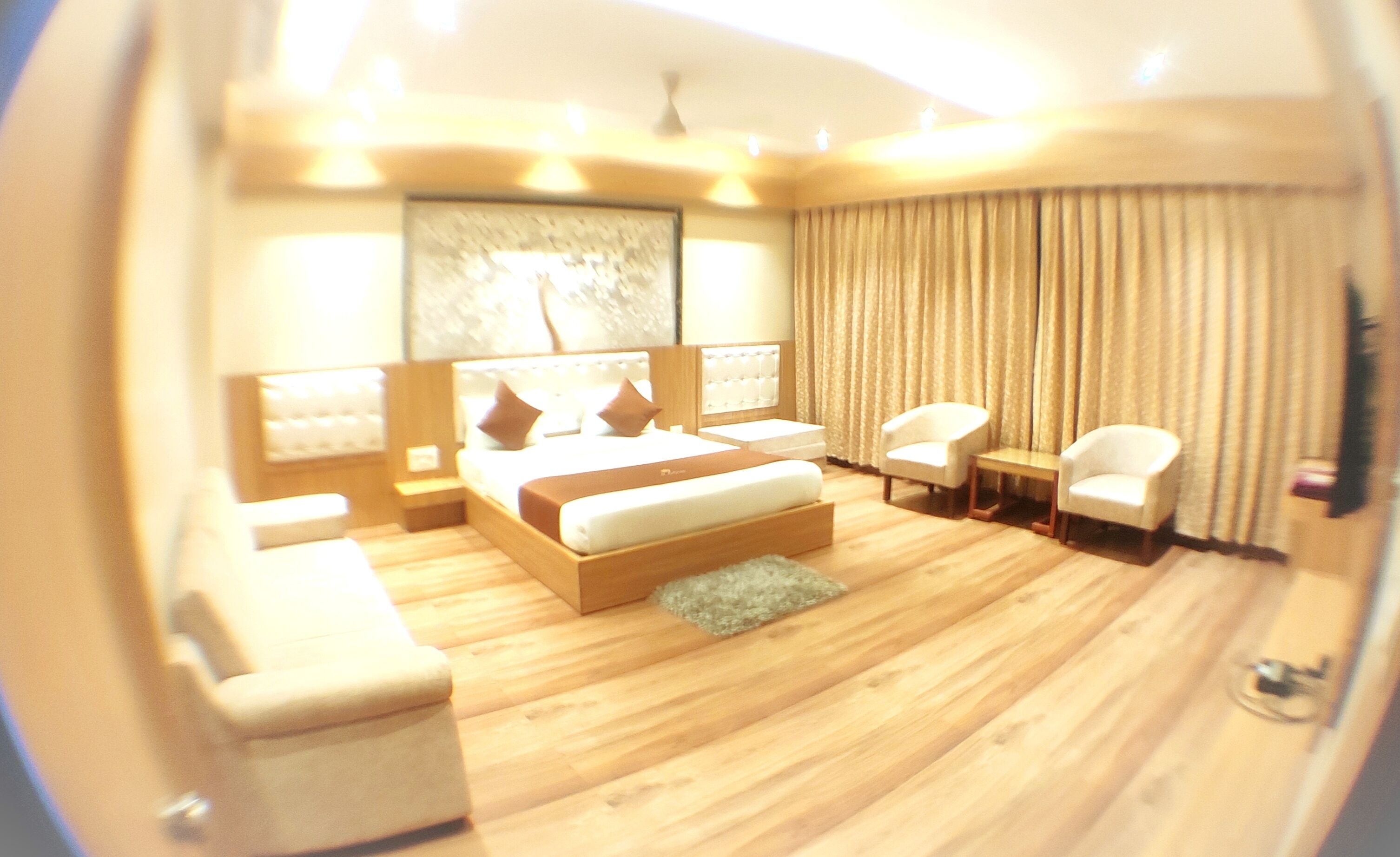 A modern hotel room with a large bed, two armchairs, a small table, and a large window with curtains.