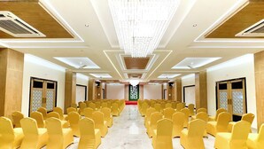 Uma sala de conferências com fileiras de cadeiras amarelas, um candelabro e um tapete vermelho que leva a um palco.