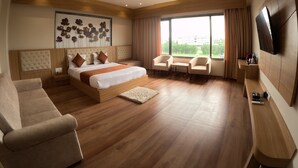 Quarto de hotel moderno com uma cama grande, duas poltronas, um sofá, uma televisão e uma janela ampla com vista para a cidade.