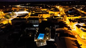 Vista nocturna de un pueblo histórico con edificios iluminados y una plaza central que tiene una fuente.