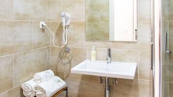 Un baño con baldosas color crema, un lavamanos blanco, un espejo, un perchero para toallas y un baúl de madera con toallas.