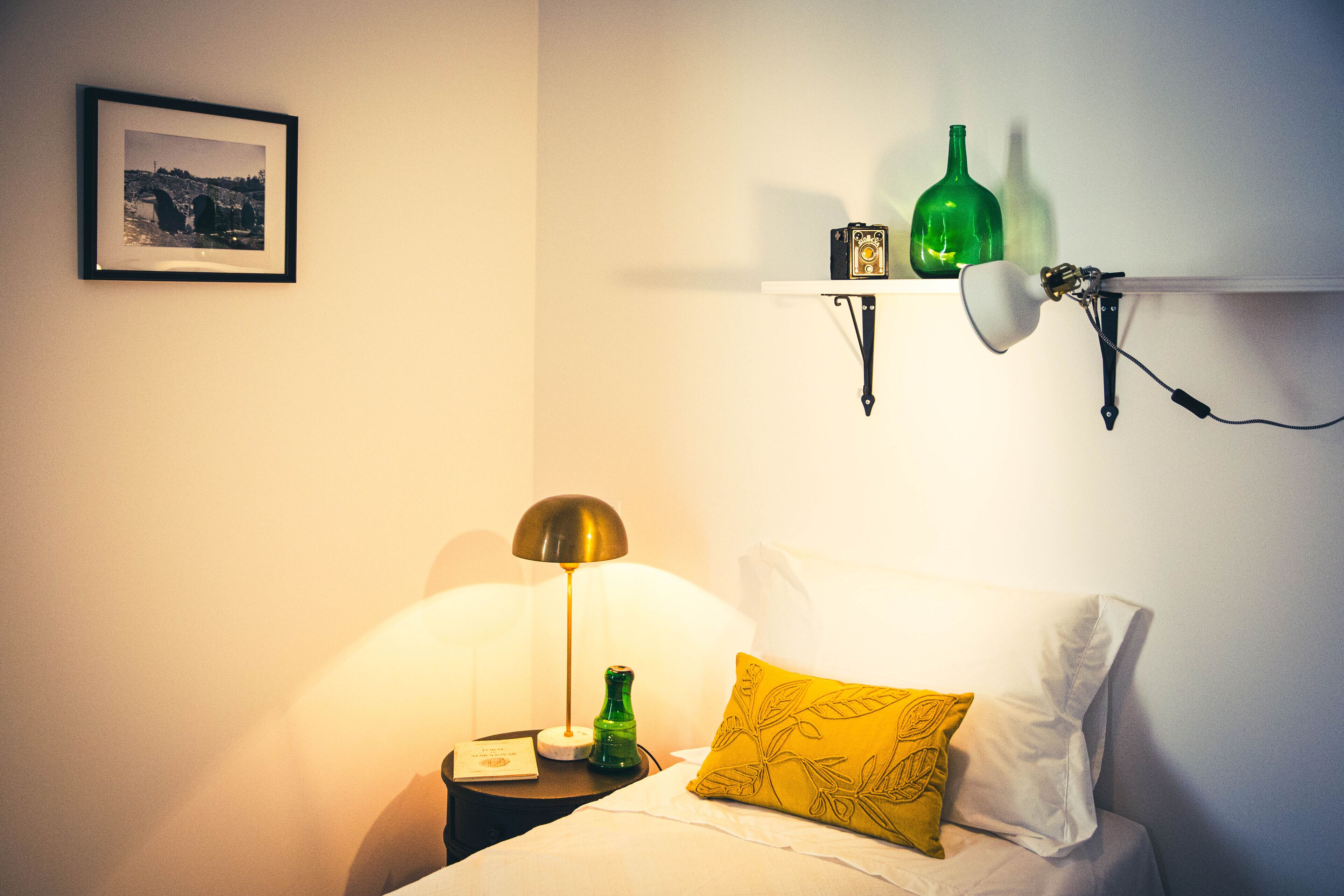 Ein ordentlich eingerichtetes Schlafzimmer mit einem Bett, einer Nachttischlampe, einem Nachttisch, einem gerahmten Bild an der Wand und einem Regal mit dekorativen Flaschen.