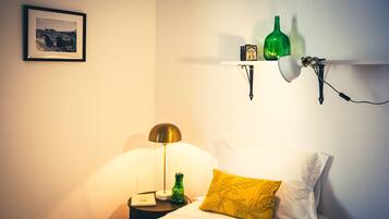 Ein ordentlich eingerichtetes Schlafzimmer mit einem Bett, einer Nachttischlampe, einem Nachttisch, einem gerahmten Bild an der Wand und einem Regal mit dekorativen Flaschen.