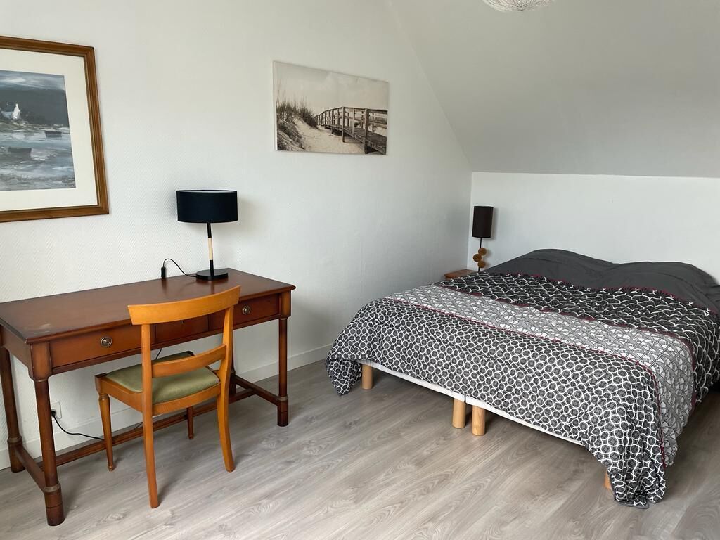 A bedroom with a wooden desk, a chair, a bed with patterned bedding, a wall-mounted lamp, and a framed picture.