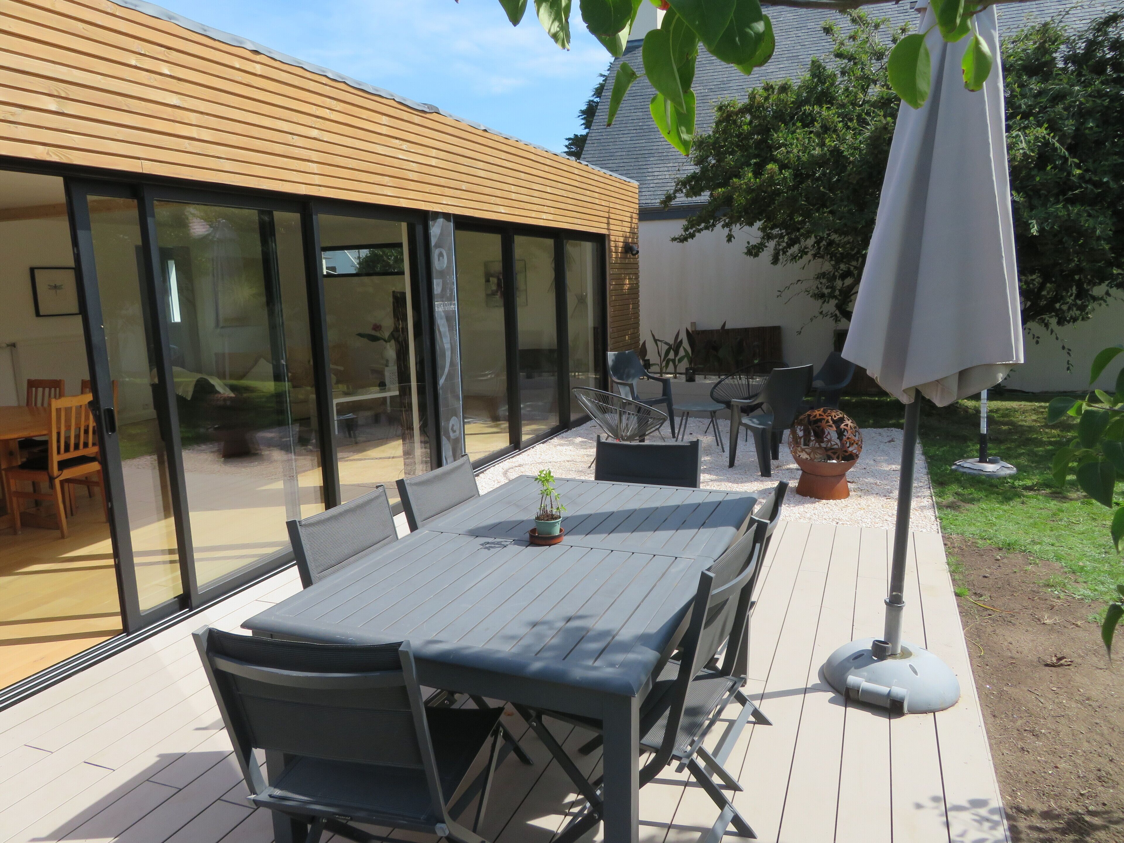 Een terras buiten met een tafel, stoelen, een bloempot en een witte parasol.