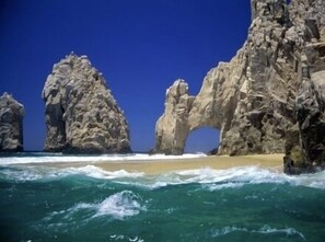 A coastal scene with large rock formations and waves crashing onto a sandy beach. - Puerta Cabos Village Oceanview 2 Bedroom (Cabo San Lucas)