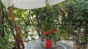 A round glass table with two white chairs, each with a red cushion, under a white umbrella. A red vase with flowers is on the table. The floor has patterned tiles. - Viareggio Sea & Garden (Viareggio)