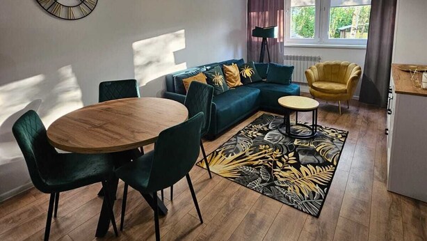 Una sala de estar moderna con una mesa de comedor de madera, sillas verdes, un sofá con cojines amarillos, una lámpara de pie y un amplio ventanal con cortinas.