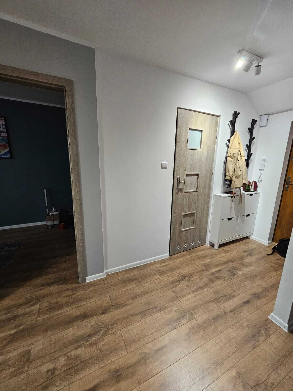 A hallway with wooden flooring, a wooden door with a small window, and a coat rack with a coat.