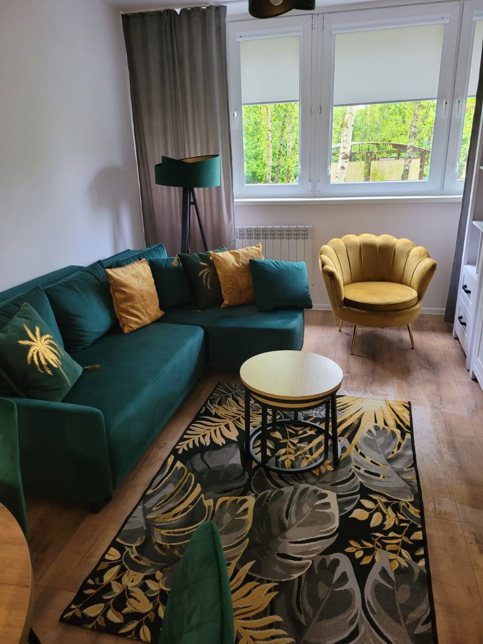 Een woonkamer met een bank, een ronde tafel, een gele fauteuil en uitzicht op groen buiten het raam.